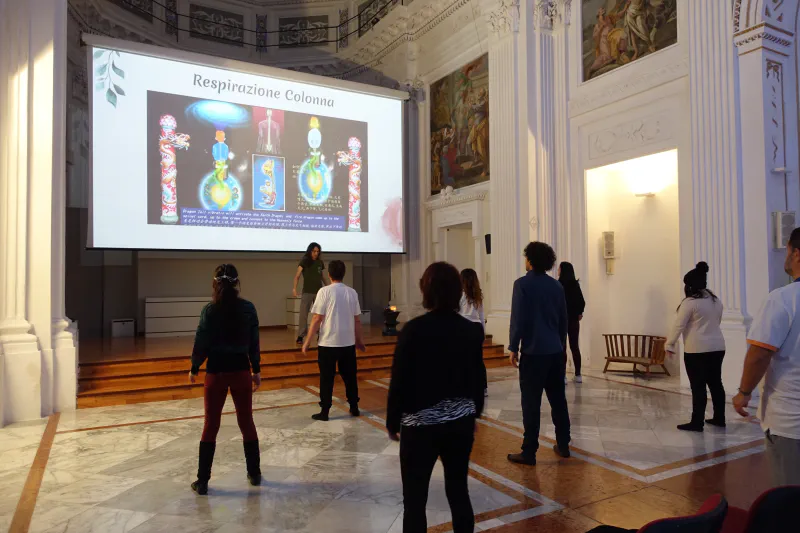 Un gruppo di partecipanti in piedi durante una lezione collettiva, in una sala storica con volte affrescate.